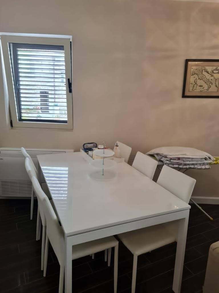 White dining set in a modern apartment living room in Petrovac, Montenegro, with a window and dark flooring.