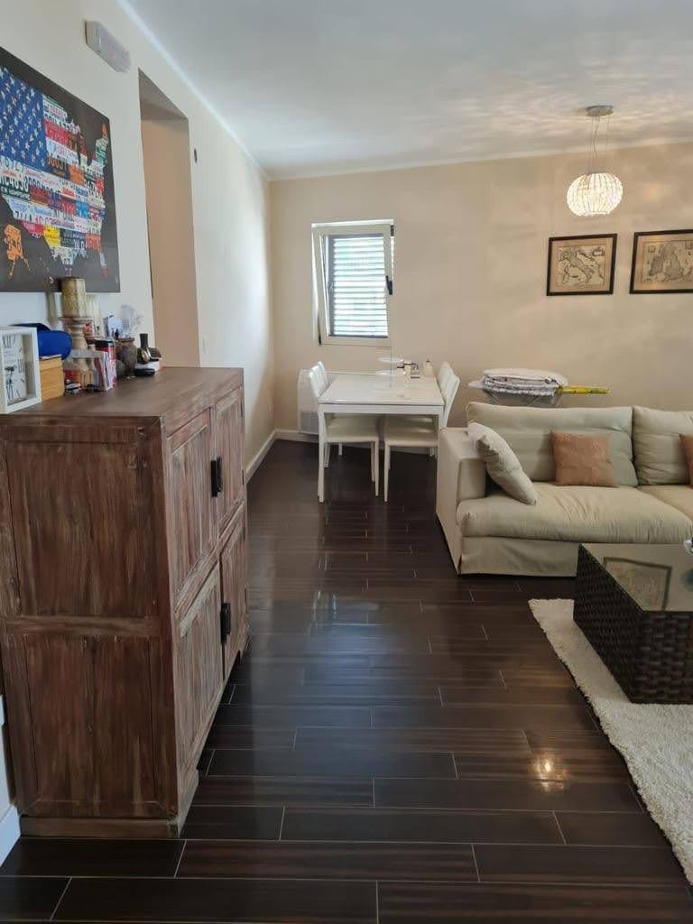 Living room in an apartment in Petrovac, Montenegro: a light sofa, dining area, wooden dresser, and dark tiled floor.