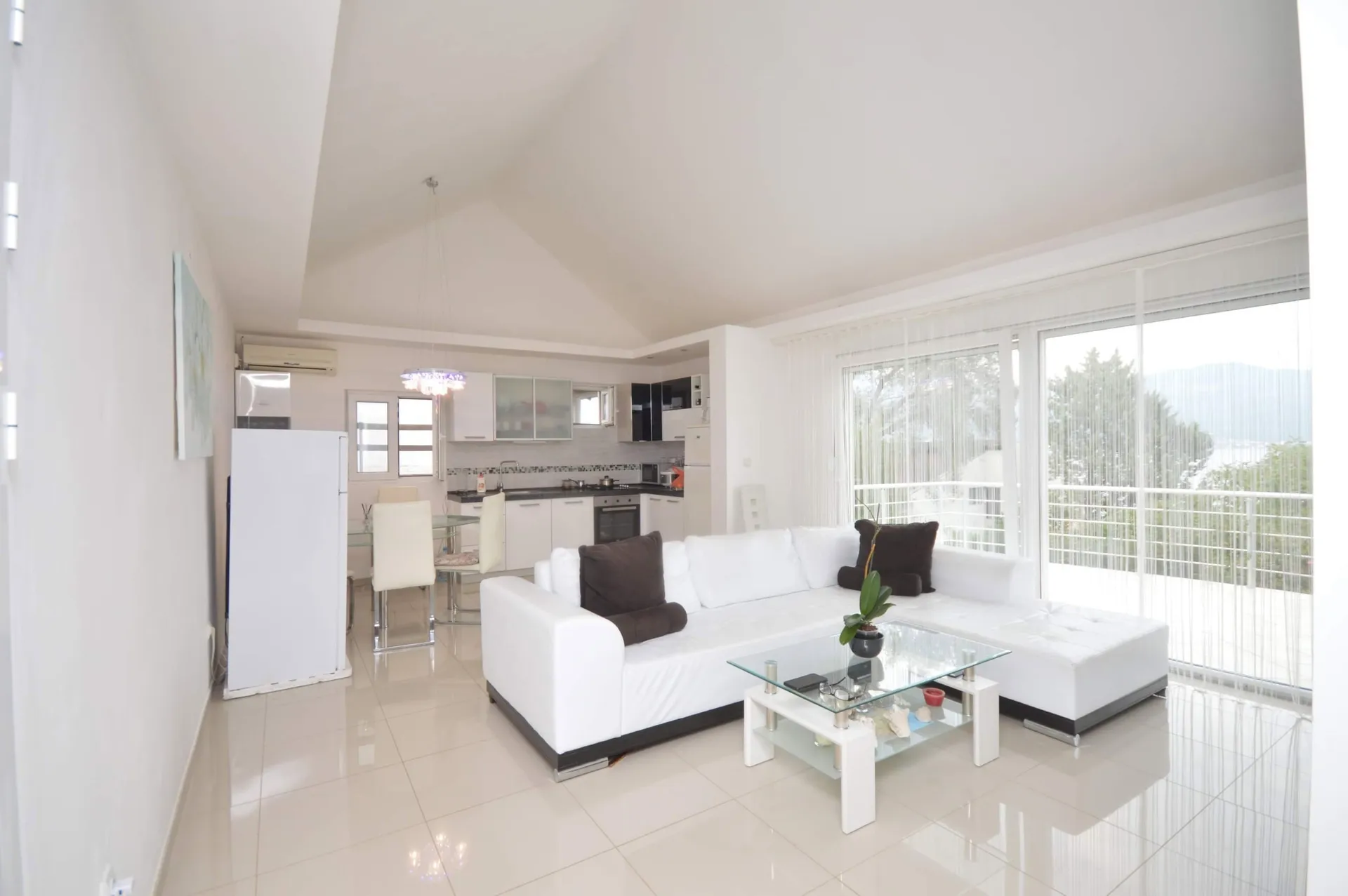 Bright living room with a white sofa, glass coffee table, and open kitchen visible in the background.