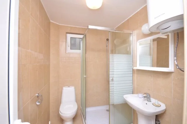 Bathroom featuring a white toilet, glass shower enclosure, pedestal sink, and wall-mounted water heater on beige tiled walls.