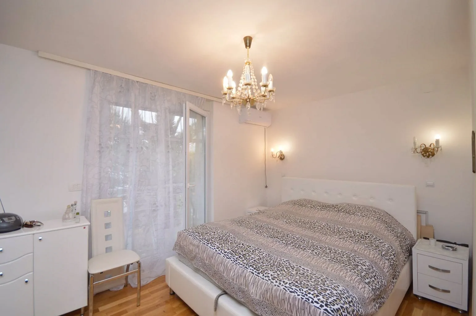 Bright bedroom featuring a white bed with leopard print bedding, crystal chandelier, and white furniture near lace curtains.