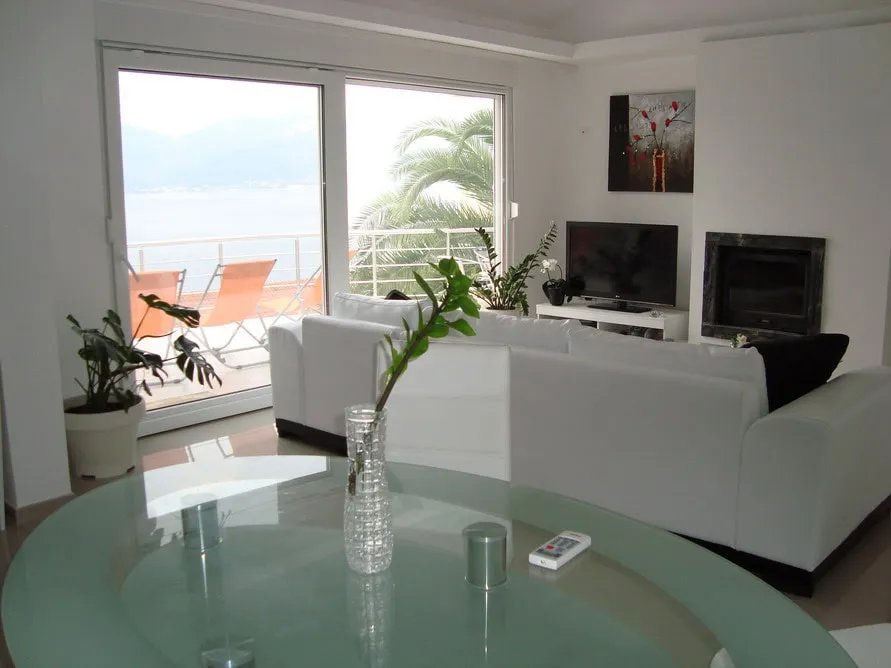 Bright living room with white sofa, glass table, TV, fireplace, and sliding doors leading to a balcony with sea view.