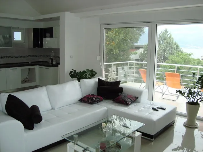 White L-shaped sofa, glass coffee table, open plan kitchen visible, large balcony doors.