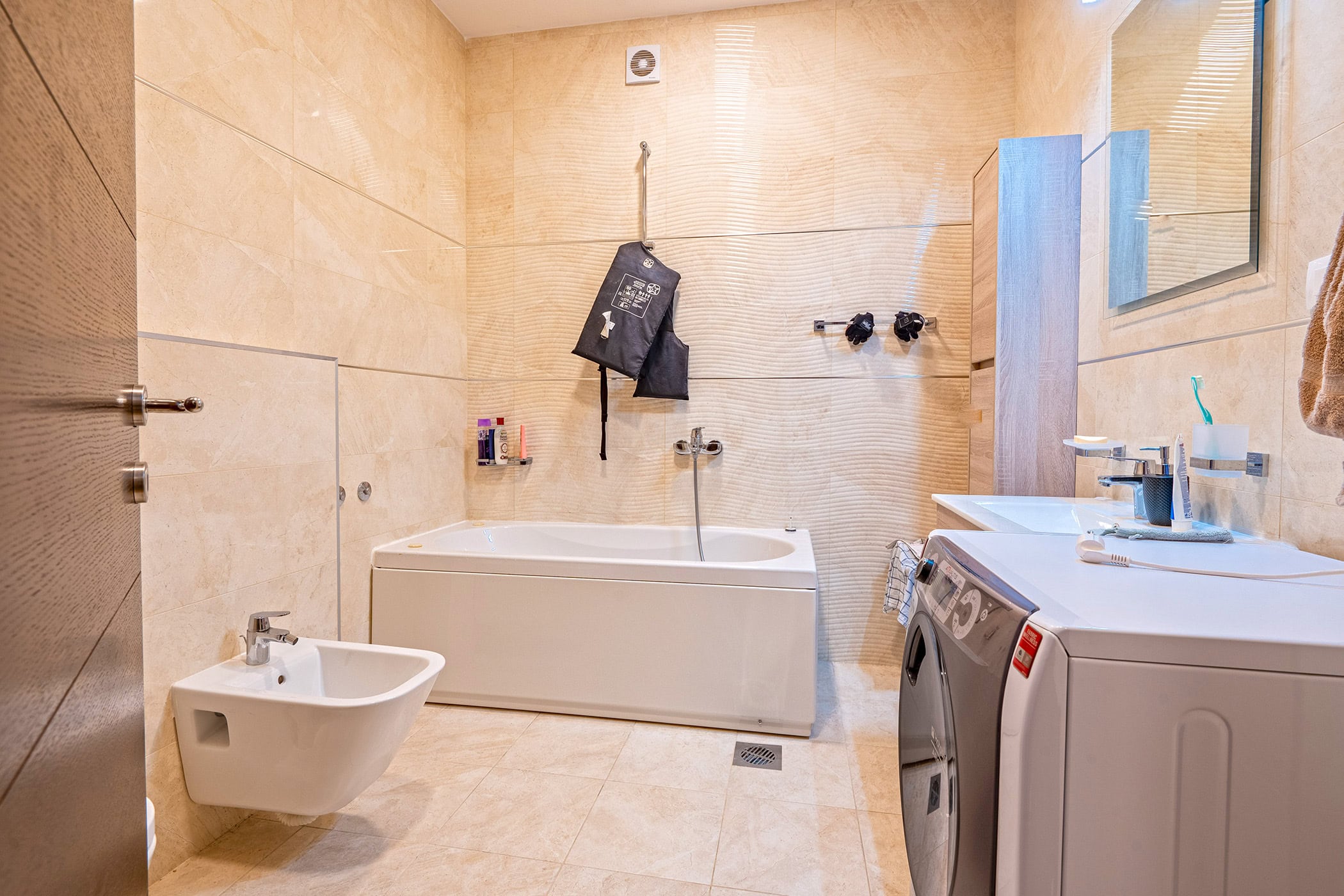 Modern bathroom with a white bathtub, bidet, and washing machine in an apartment in Budva, Montenegro. Beige tiles.