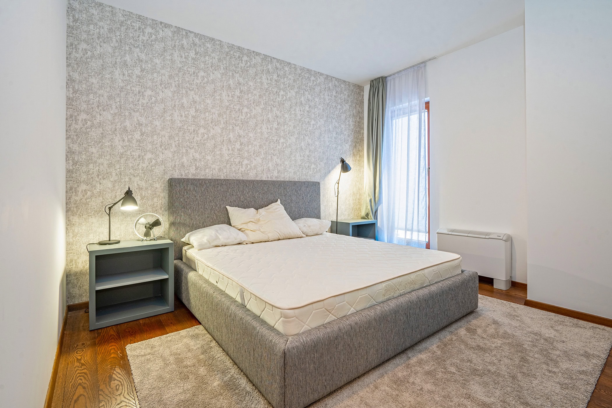 Cozy bedroom with a double bed in an apartment in Budva, Montenegro: gray wallpaper, parquet flooring, and access to the balcony.