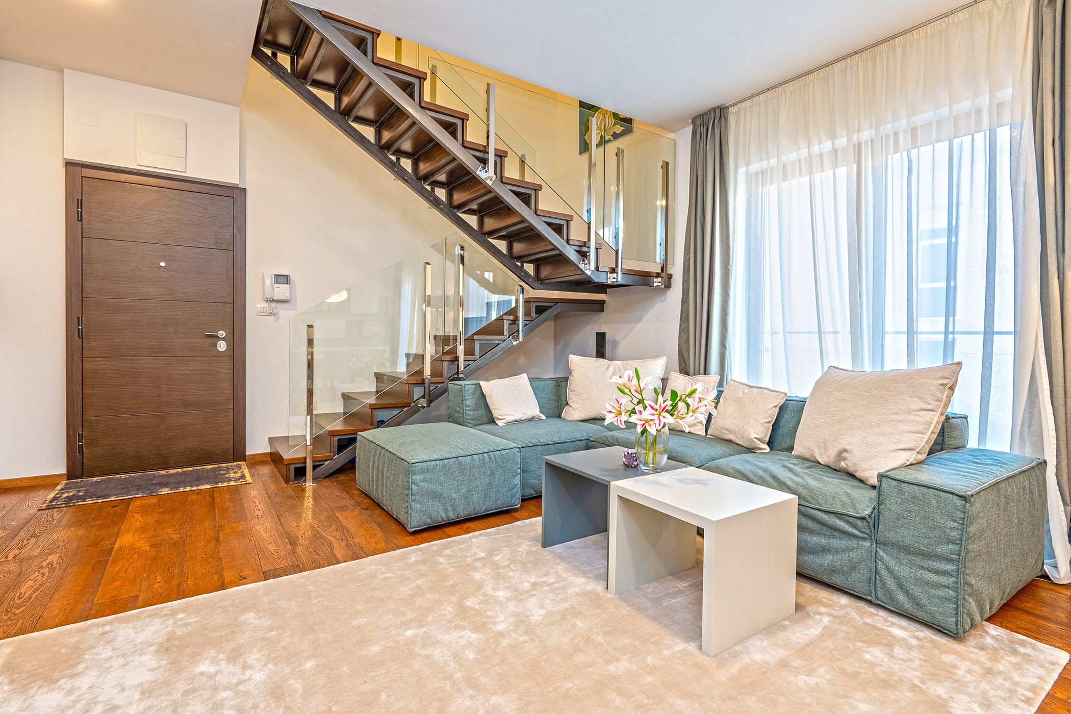 Living room in a duplex in Budva, Montenegro: modern staircase with glass railing, sofa, and parquet floor.