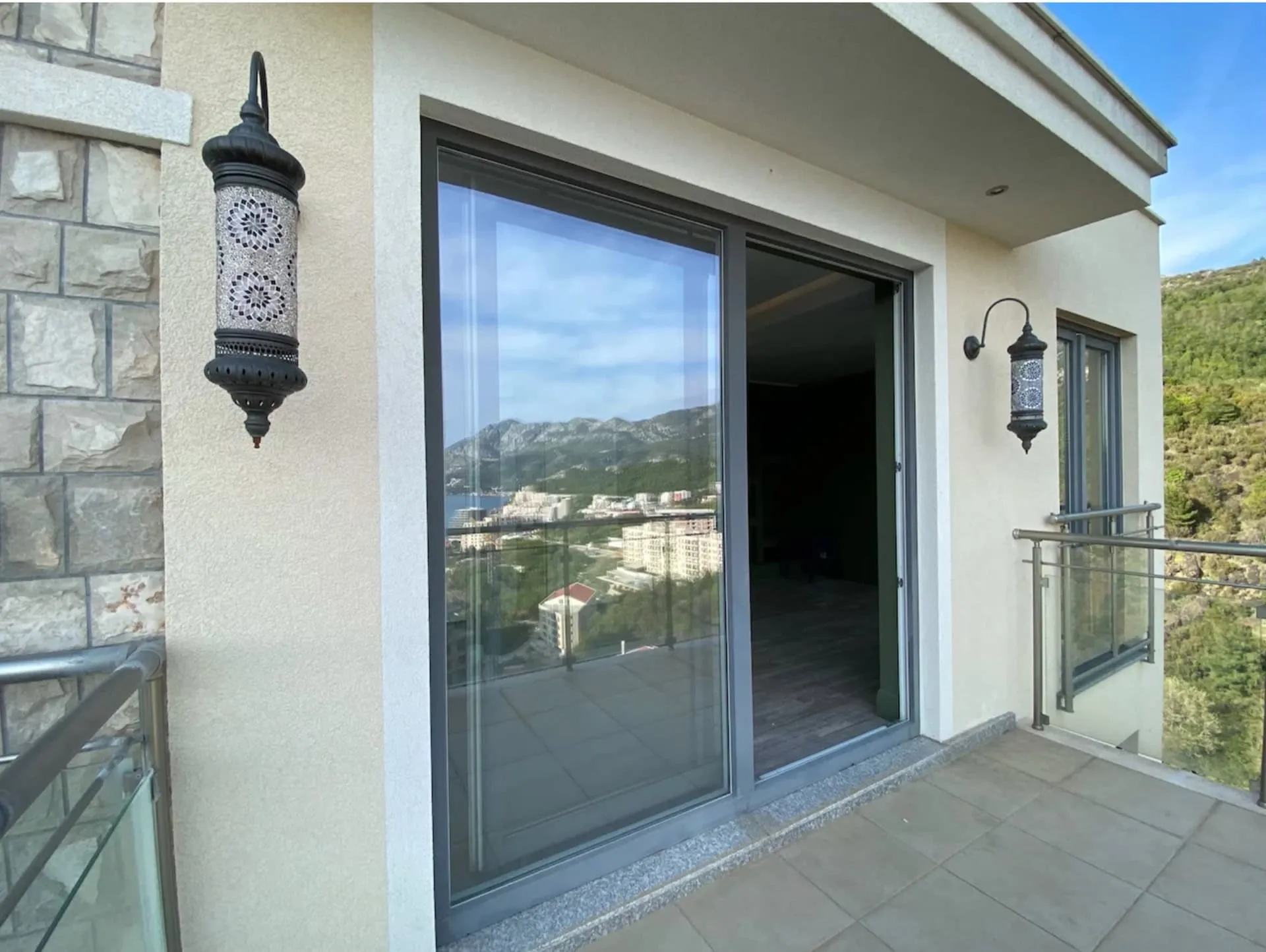 Balcony with glass railing, sliding door, and decorative wall lanterns overlooking a city and mountains.
