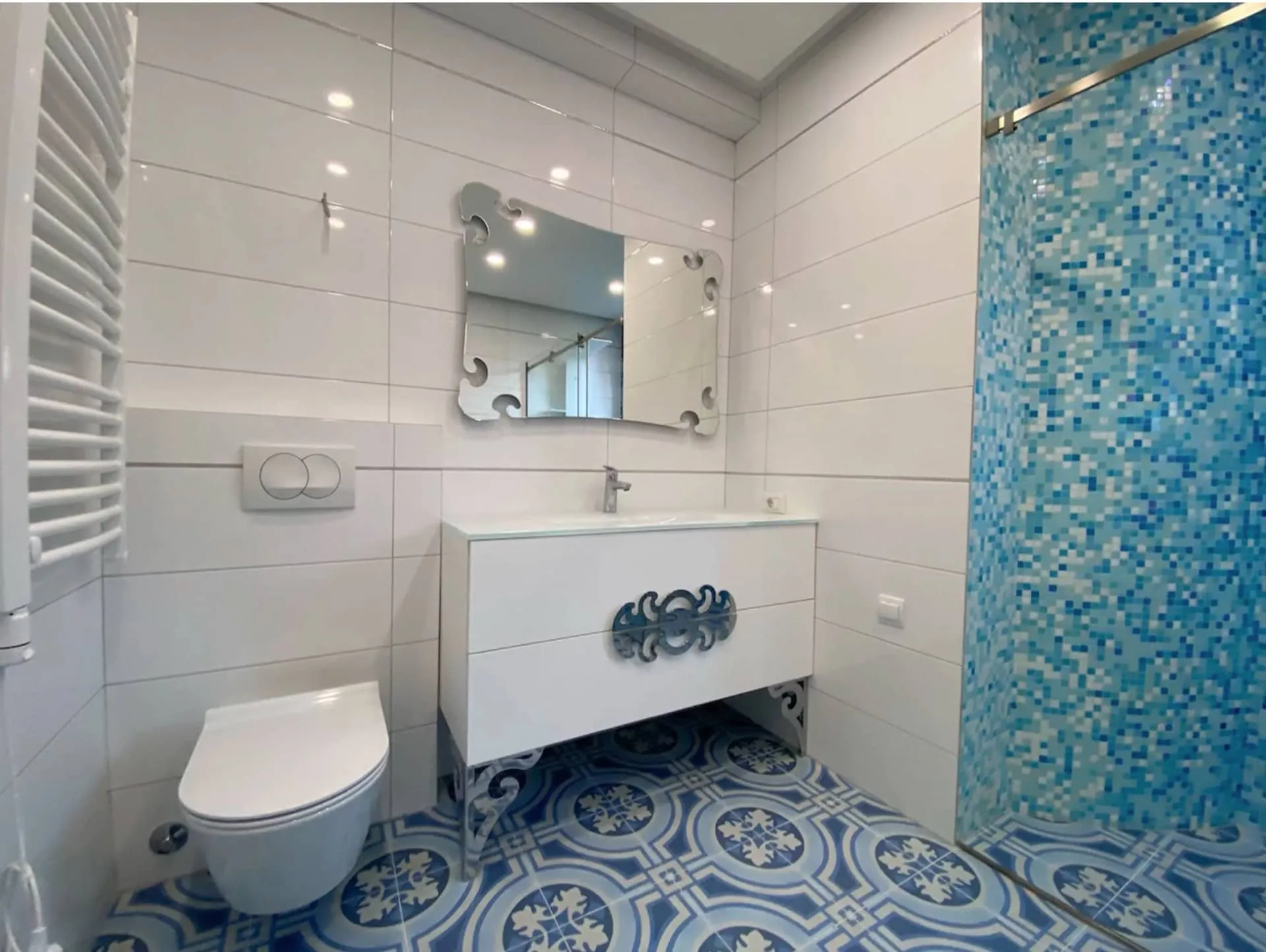 Bathroom with white toilet, vanity, and mirror. Features blue shower mosaic and patterned floor tiles.