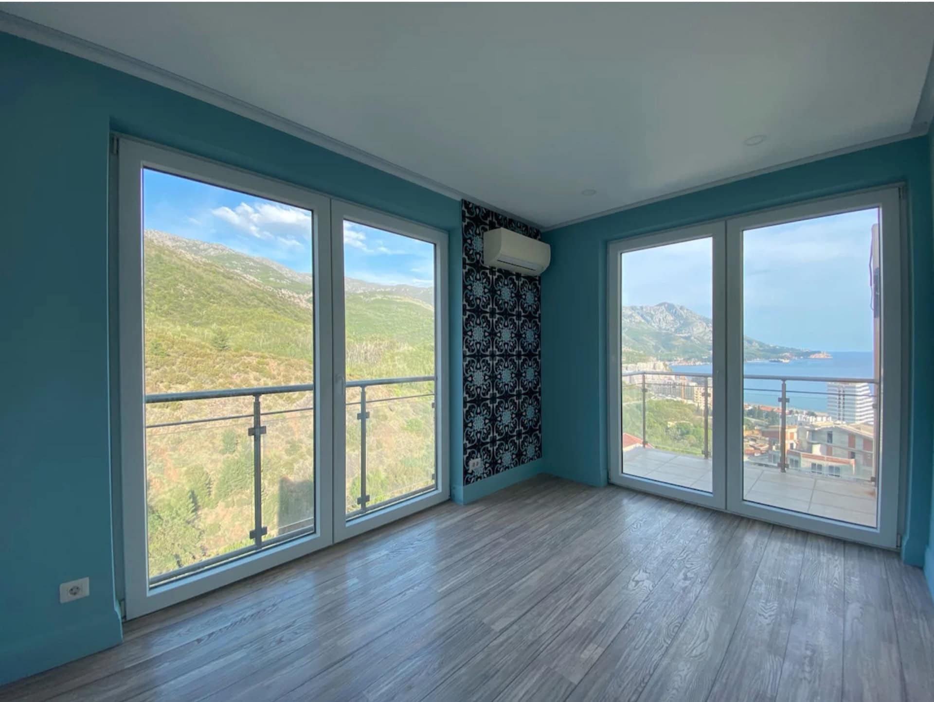 Living room in an apartment in Bečići, Montenegro: panoramic windows with sea and mountain views, laminate flooring, and an accent wall.
