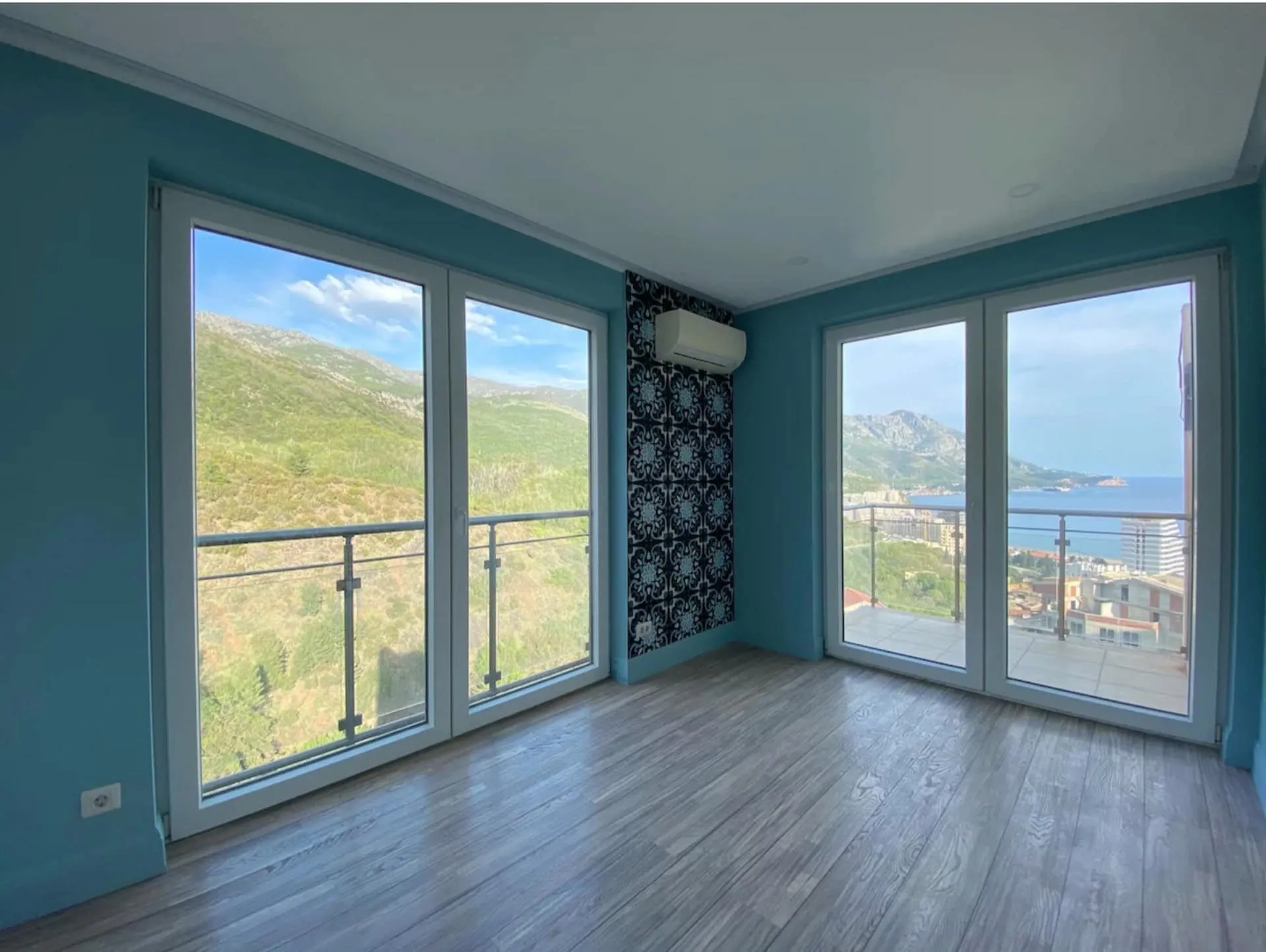 Empty room with teal walls, wood flooring, and large glass doors offering a panoramic view of the sea and mountains.