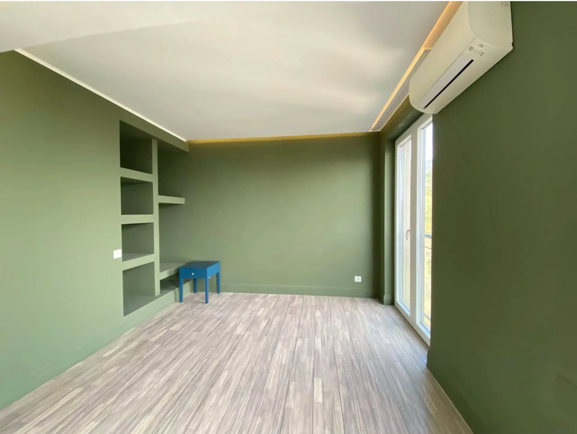 Empty room with green walls, built-in shelves, a small blue table, and a wall-mounted AC unit.