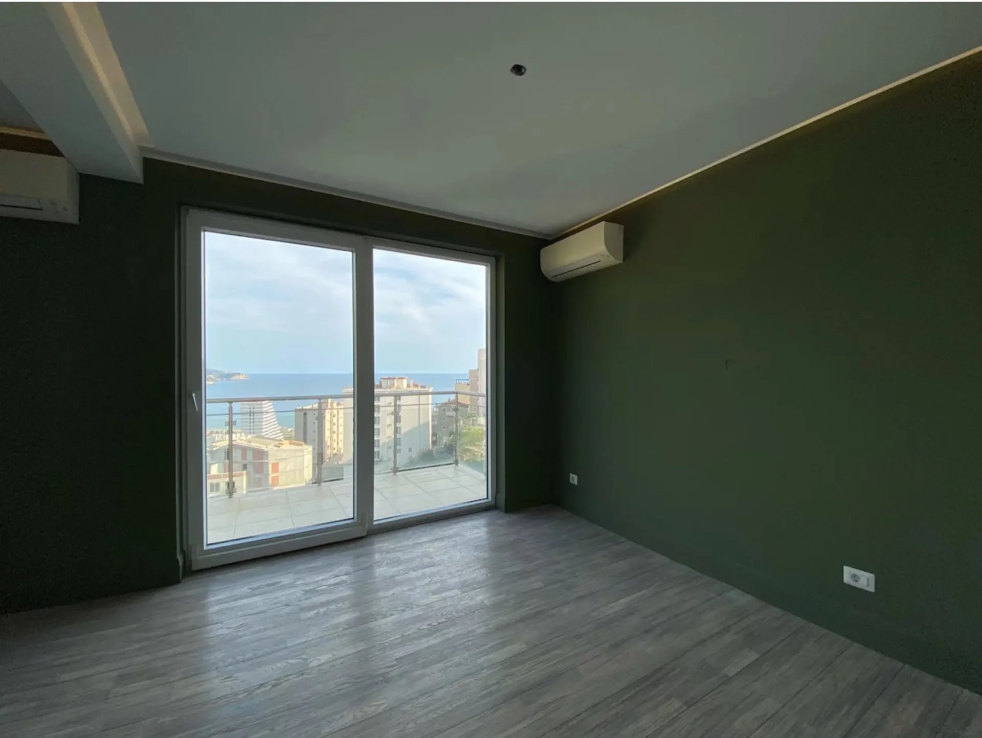 Empty room with dark green walls, wood laminate flooring, and a large sliding glass door leading to a balcony with a sea view.