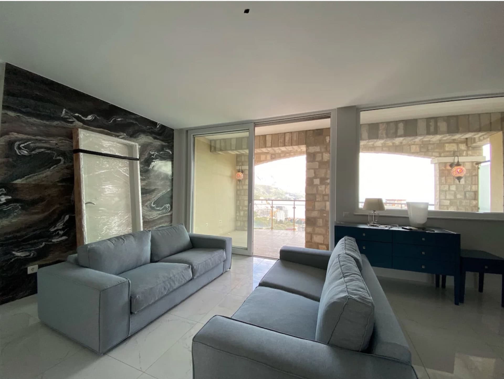 Living room in an apartment in Bečići, Montenegro: gray sofas, a stone accent wall, and access to a terrace with sea views.