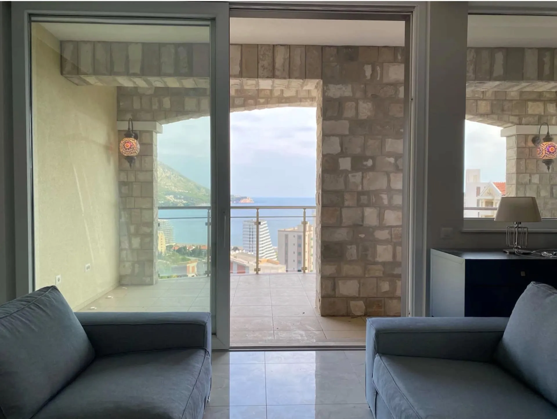 Living room with grey sofas opening to a stone-arched balcony overlooking the sea and city.