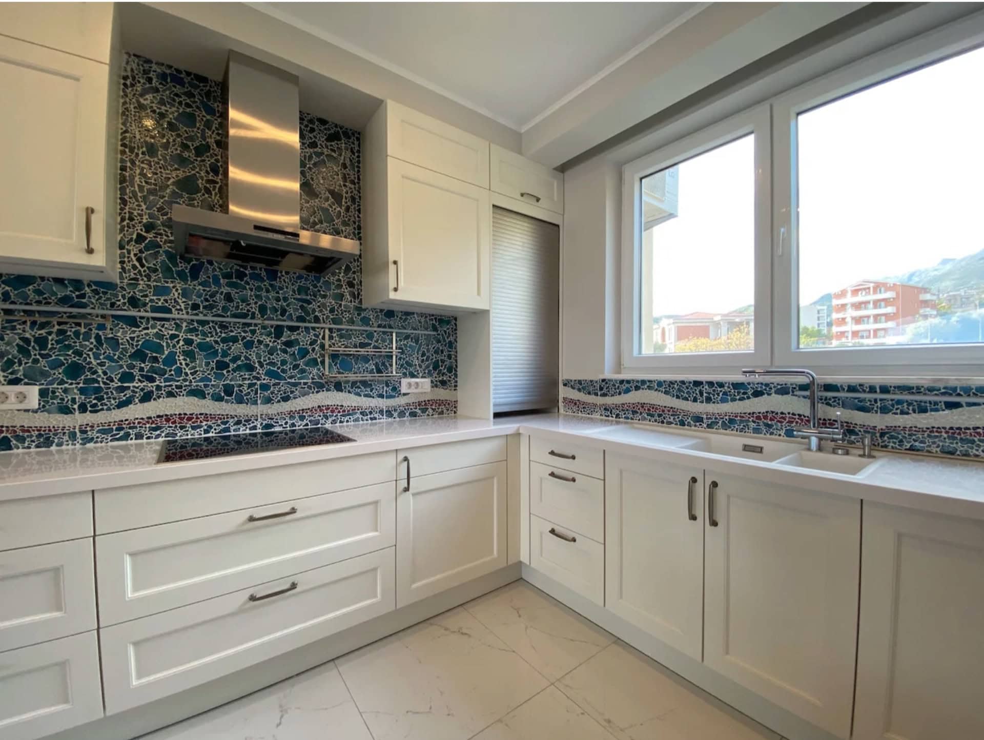 Modern white kitchen with a mosaic backsplash in an apartment, Bečići, Montenegro. Corner kitchen set and a view from the window.