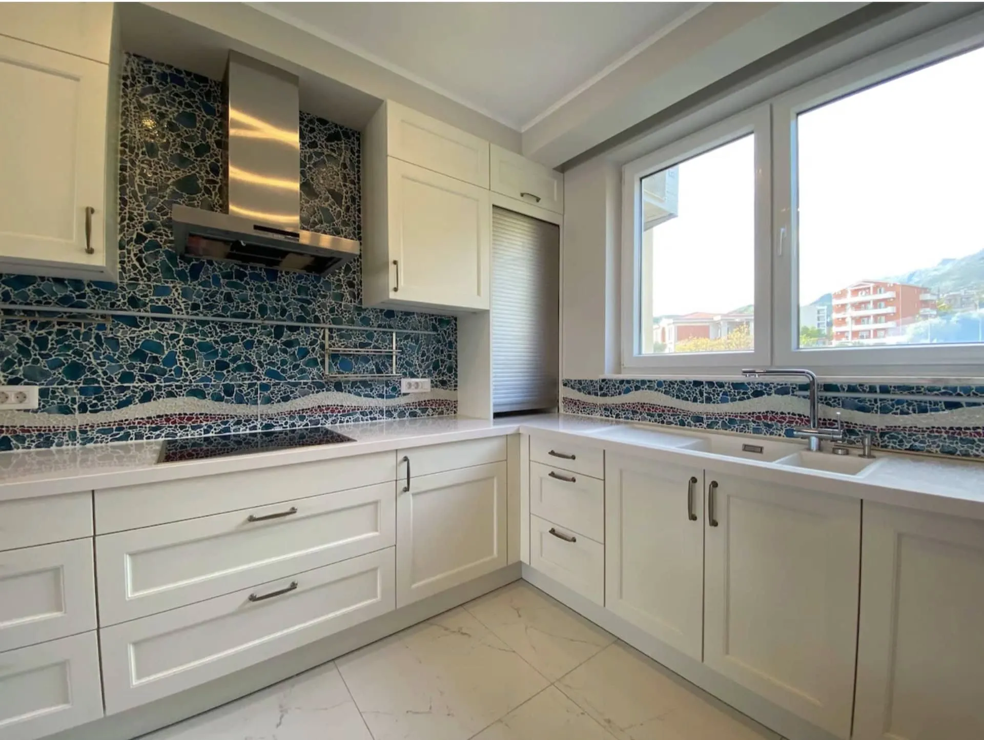Modern kitchen featuring white cabinetry, a blue mosaic backsplash, induction cooktop, and sink near a window.