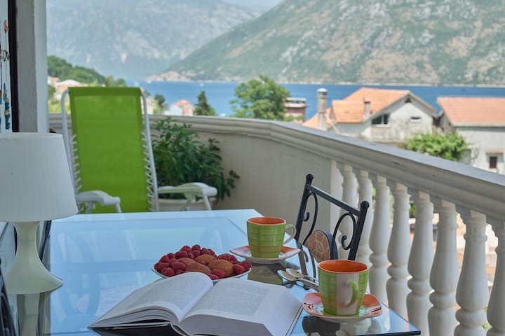 Balcony with a view of Kotor Bay in an apartment, Prčanj, Montenegro: glass table, cups, berries, and a sun lounger.