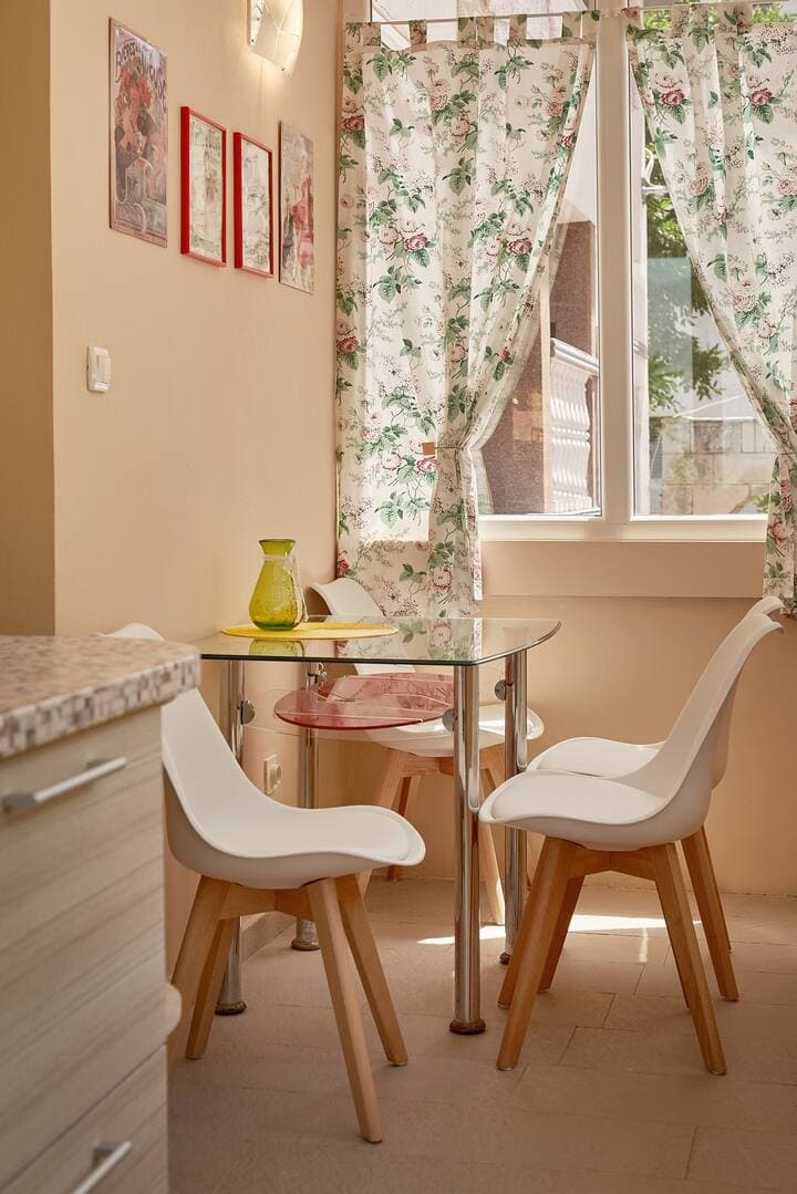 Cozy dining area in the kitchen of an apartment in Prčanj, Montenegro: a glass table and white chairs by the window with curtains.