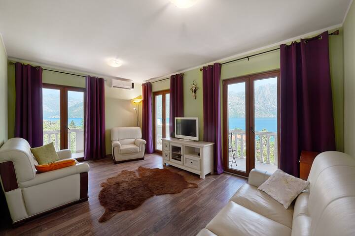 Living room in an apartment in Prčanj, Montenegro: light furniture, a rug, and panoramic windows with sea and mountain views.