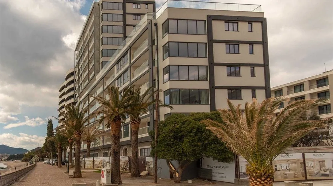 Exterior view of a multi-story residential building with balconies and palm trees along a promenade.