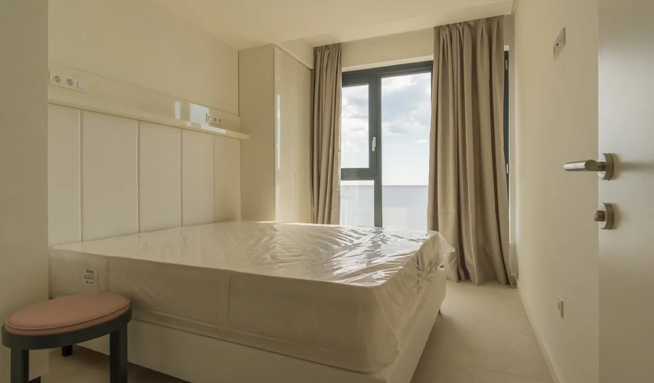 Bedroom with plastic-wrapped mattress, white headboard, wardrobe, and sea-view window.