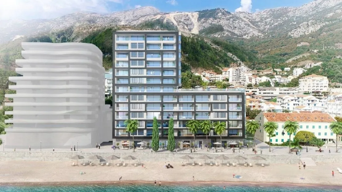 Exterior view of a modern multi-story seaside apartment building with a beach and mountains in the background.