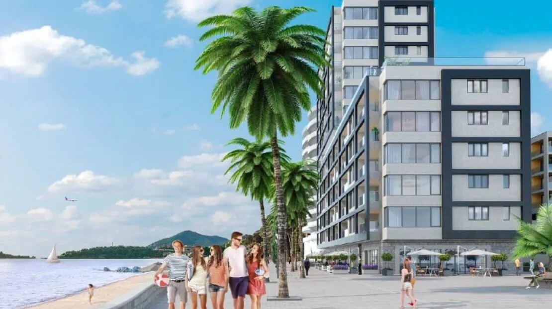 Exterior view of a modern multi-story seaside building with palm trees and people walking on the promenade.