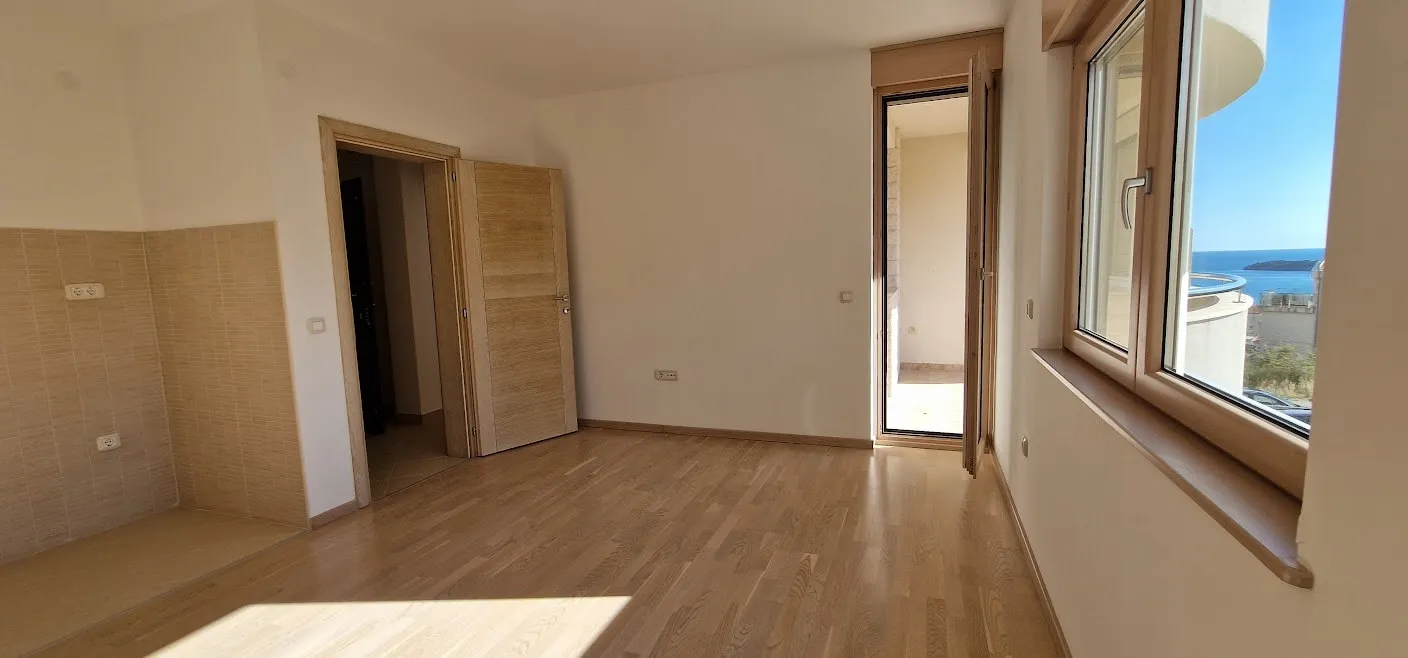 Empty room with laminate flooring and a tiled wall section, featuring a door and window overlooking the sea.