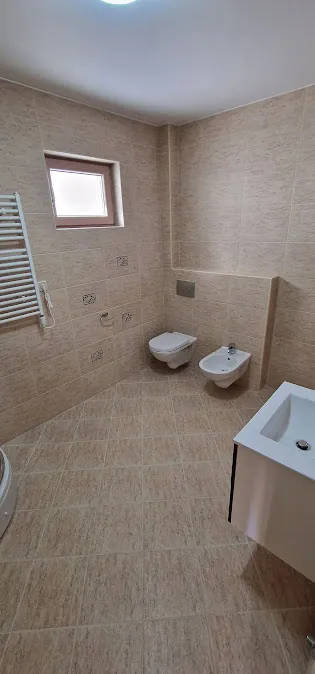 Bathroom with beige tiles, wall-mounted toilet, bidet, and partial view of a sink vanity.