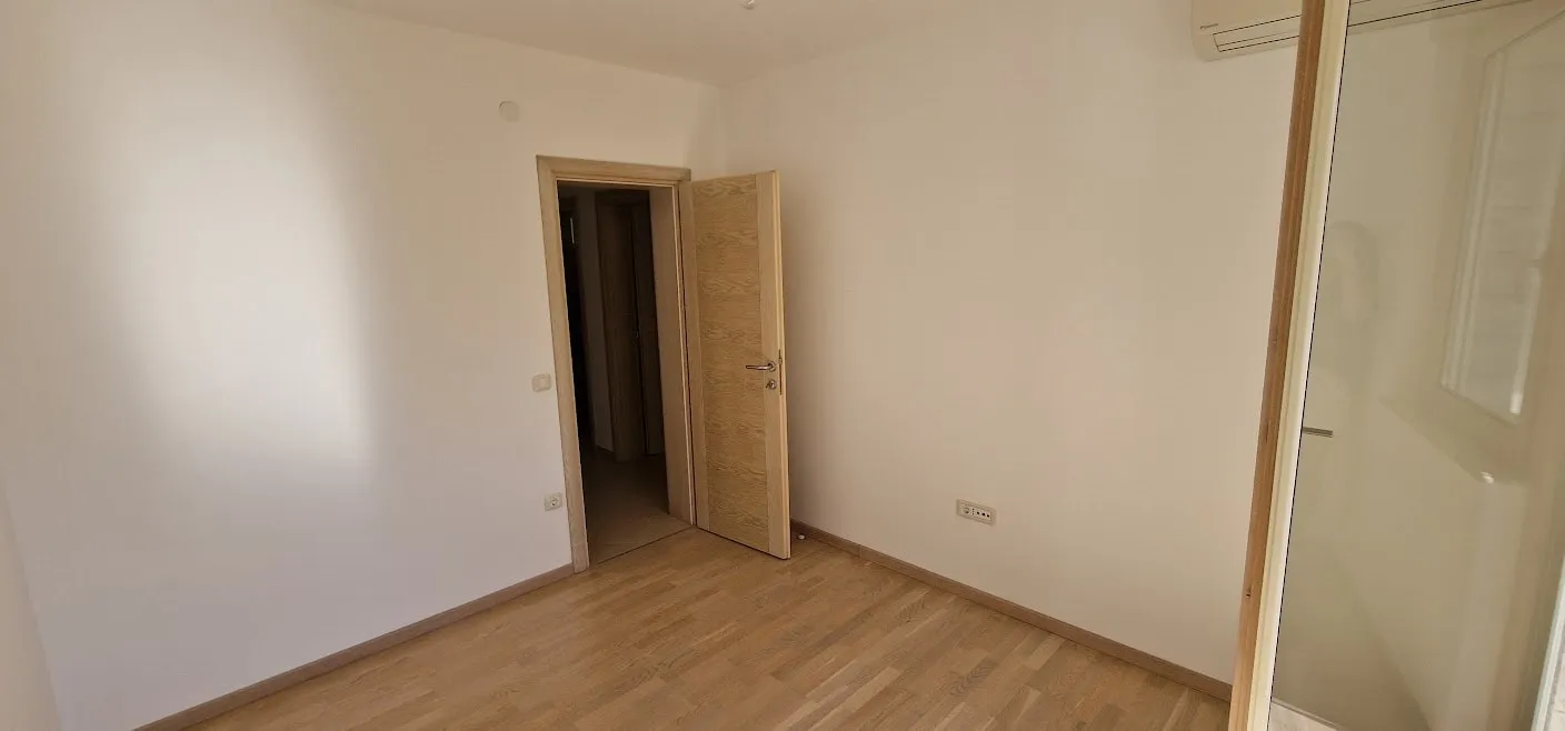 Empty room with laminate flooring and white walls. An open wooden door leads to a hallway, and an AC unit is visible.