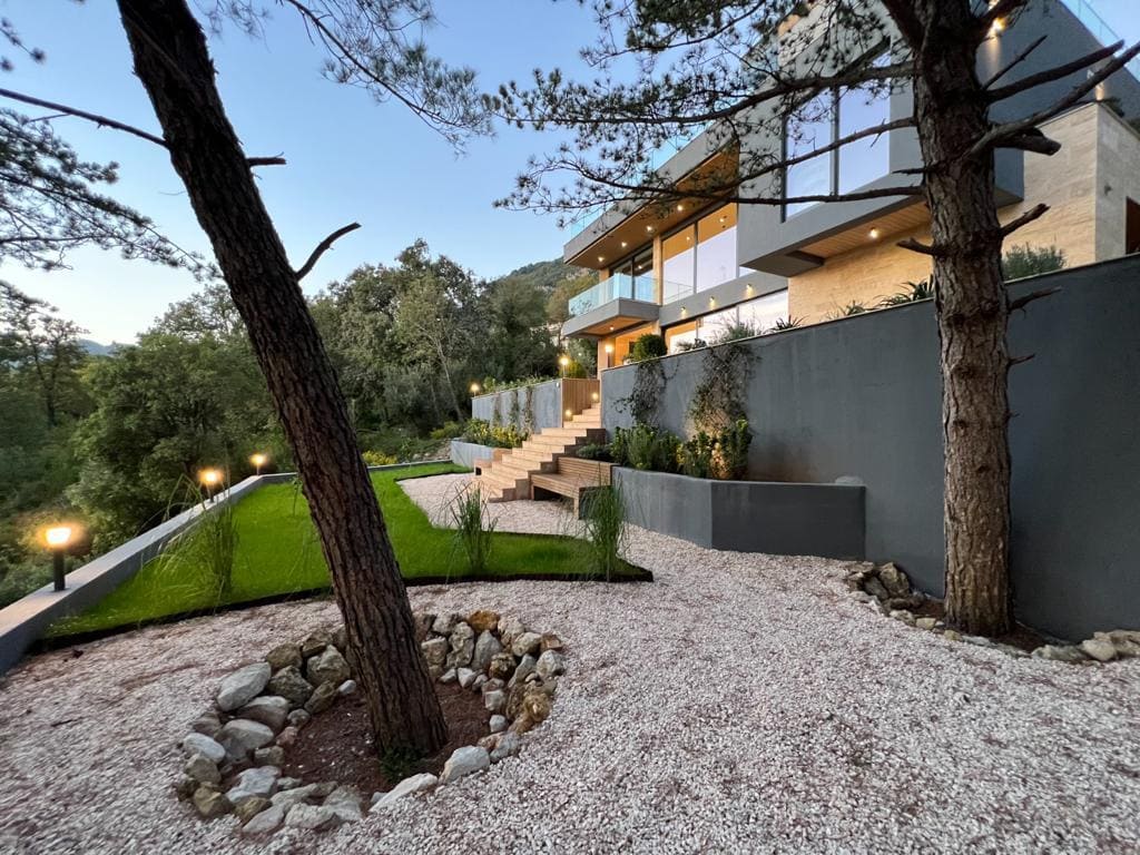 Modern villa in Kavac, Montenegro: panoramic glazing, landscape design, terrace, and garden with pine trees.