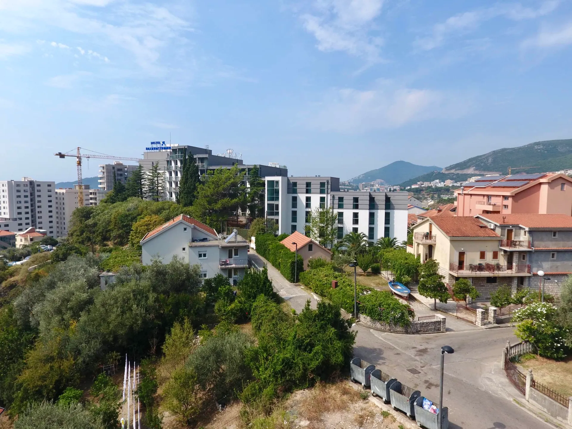 Panoramic exterior view of a town featuring residential buildings, a hotel, lush greenery, and mountains.