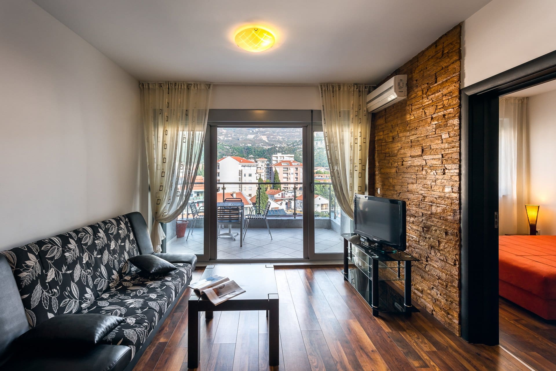 Living room in an apartment in Budva, Montenegro: sofa, stone finish, TV, and access to a balcony with city and mountain views.
