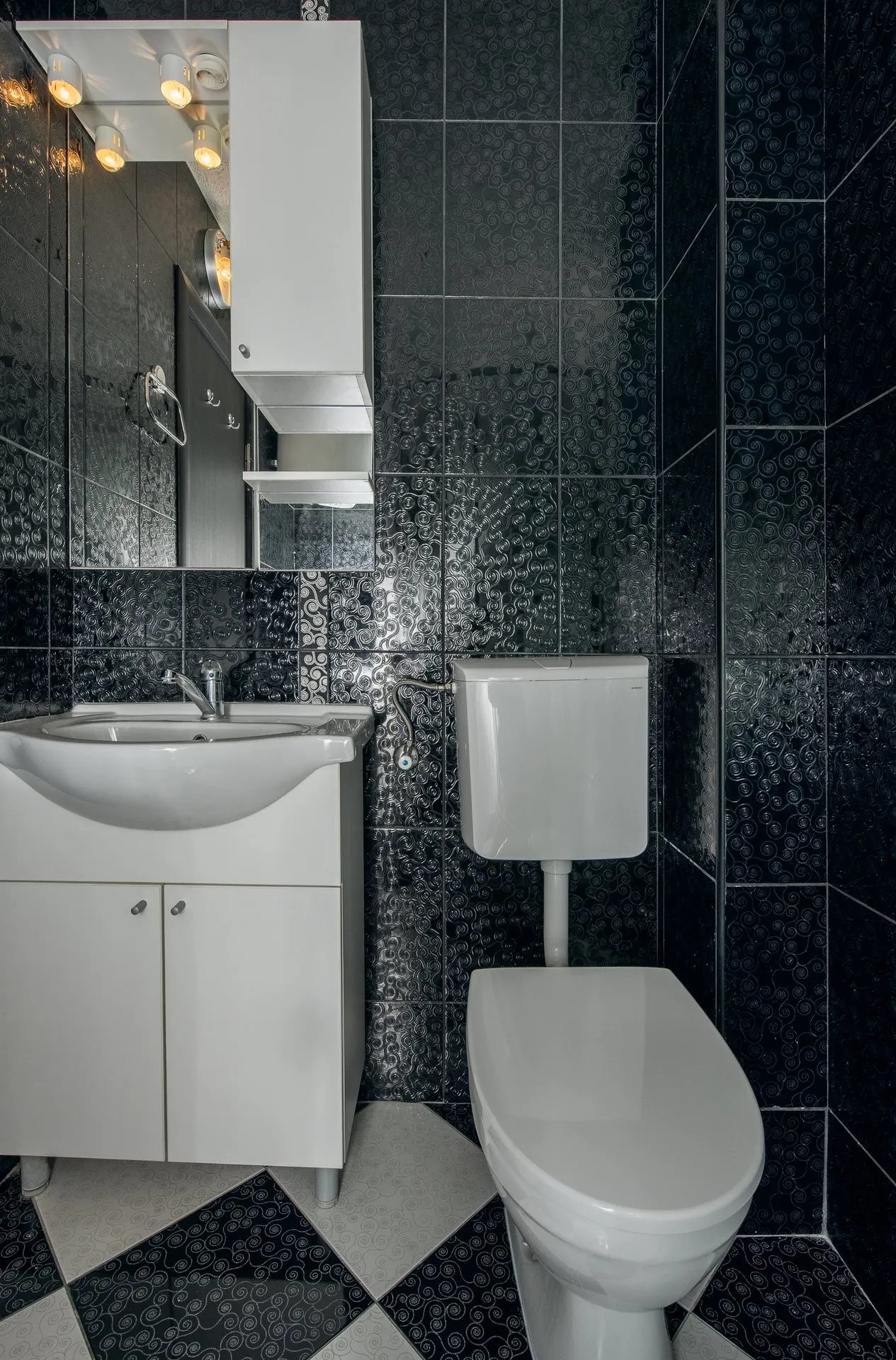 Bathroom with black textured tiles, white toilet, vanity sink, and a mirror cabinet equipped with warm lighting.