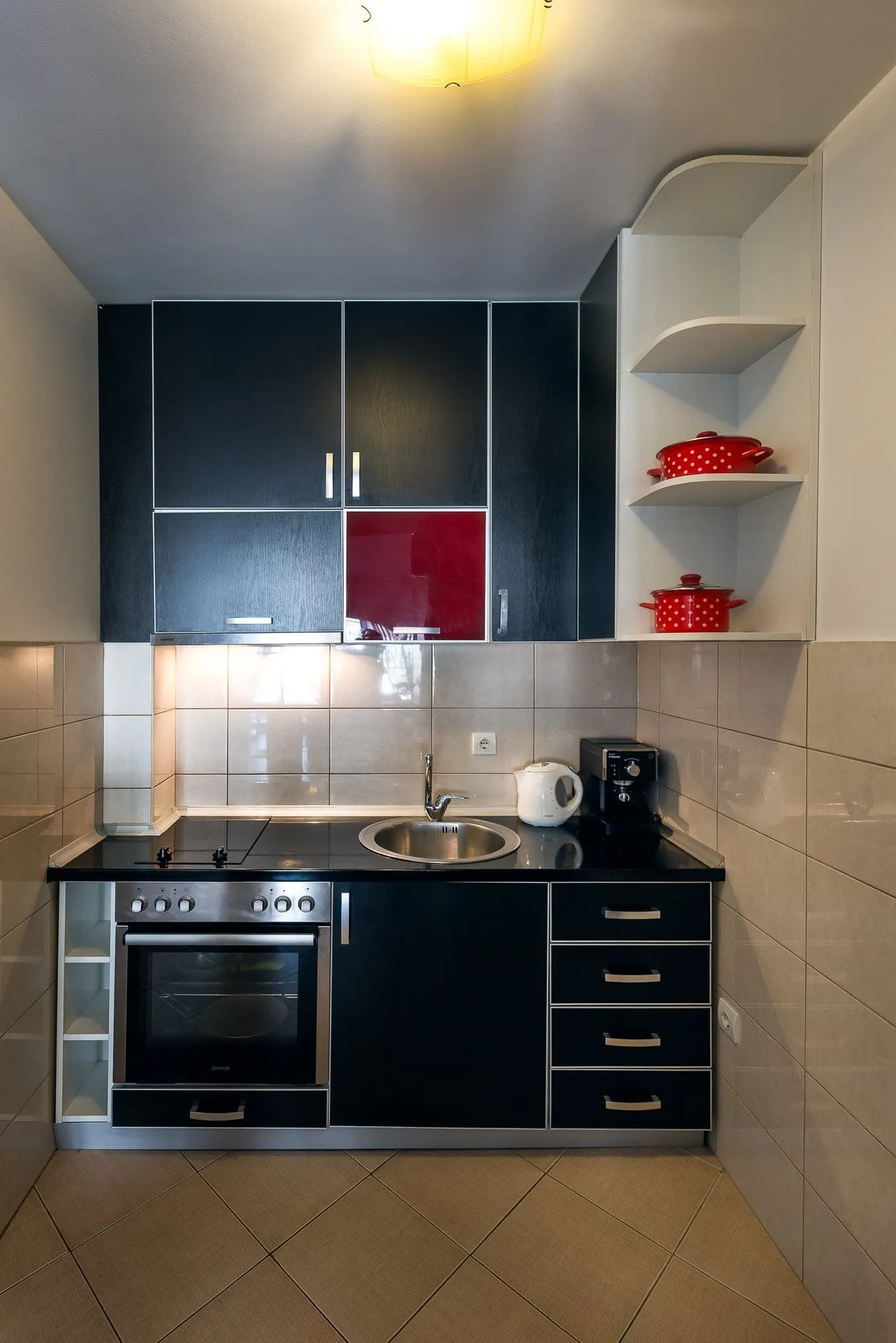 Compact kitchen with black cabinets, a red accent panel, stove, sink, and appliances like a kettle and coffee maker on the counter.