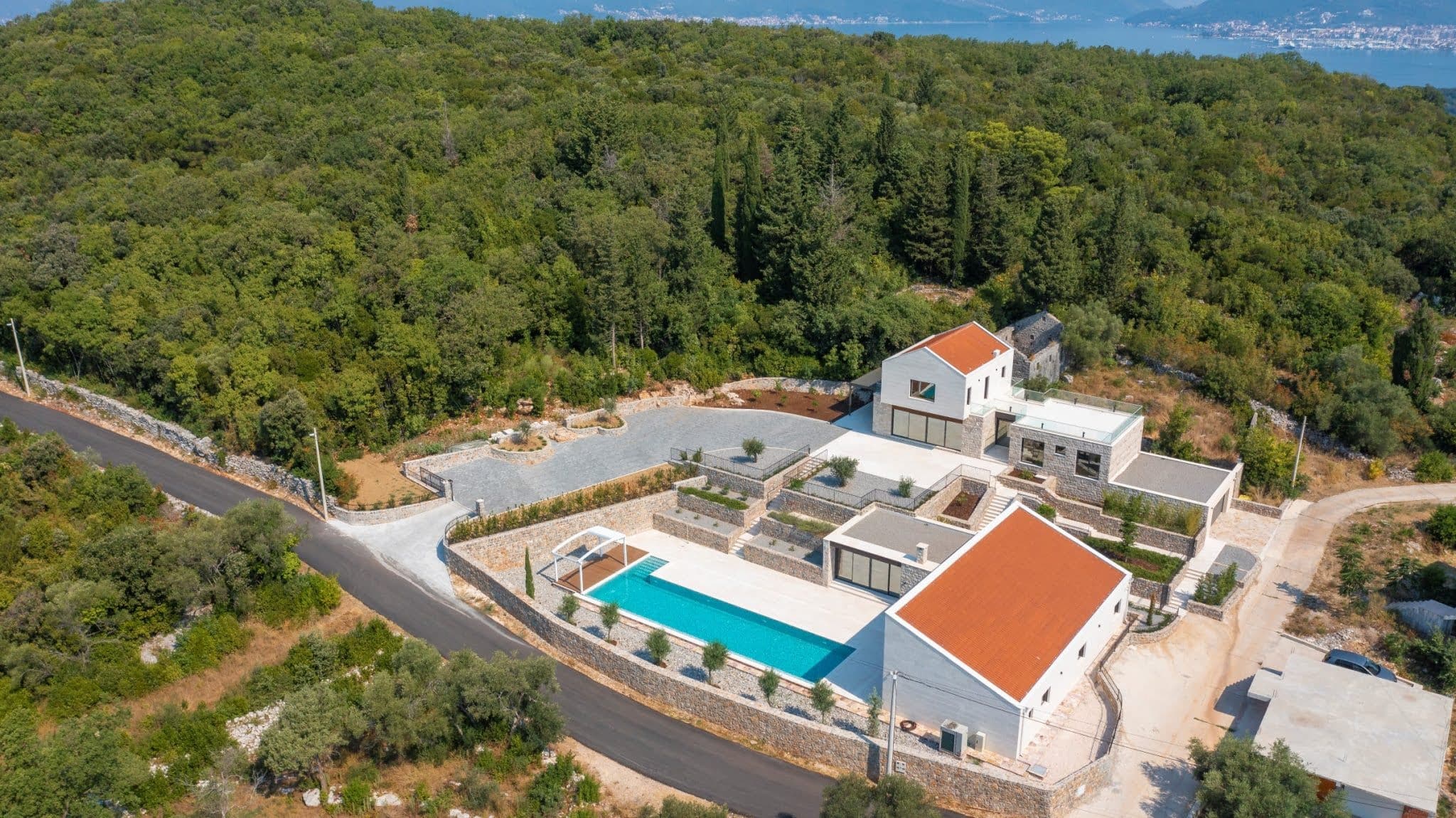 Aerial view of a villa complex with a pool and terraces in Tivat, Montenegro, against a backdrop of forest and coastline.