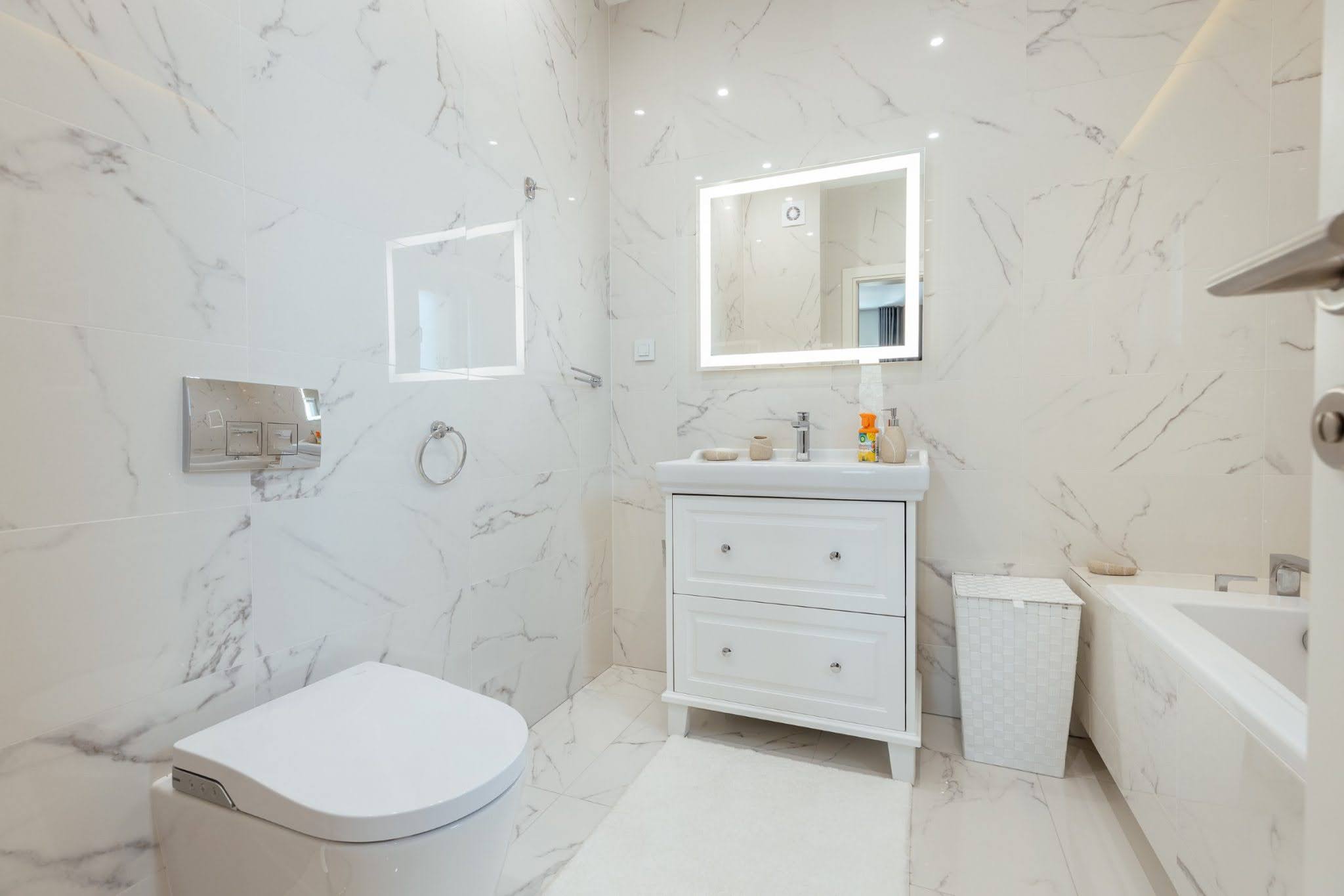 Modern bathroom with white marble finish in Kumbor, Montenegro: vanity, backlit mirror, and bathtub.
