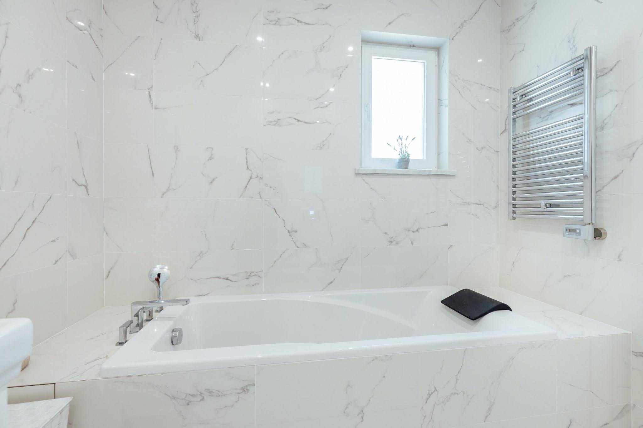 Modern bathroom in Kumbor residential complex, Montenegro: white marble-look tiles, built-in bathtub, and towel warmer.