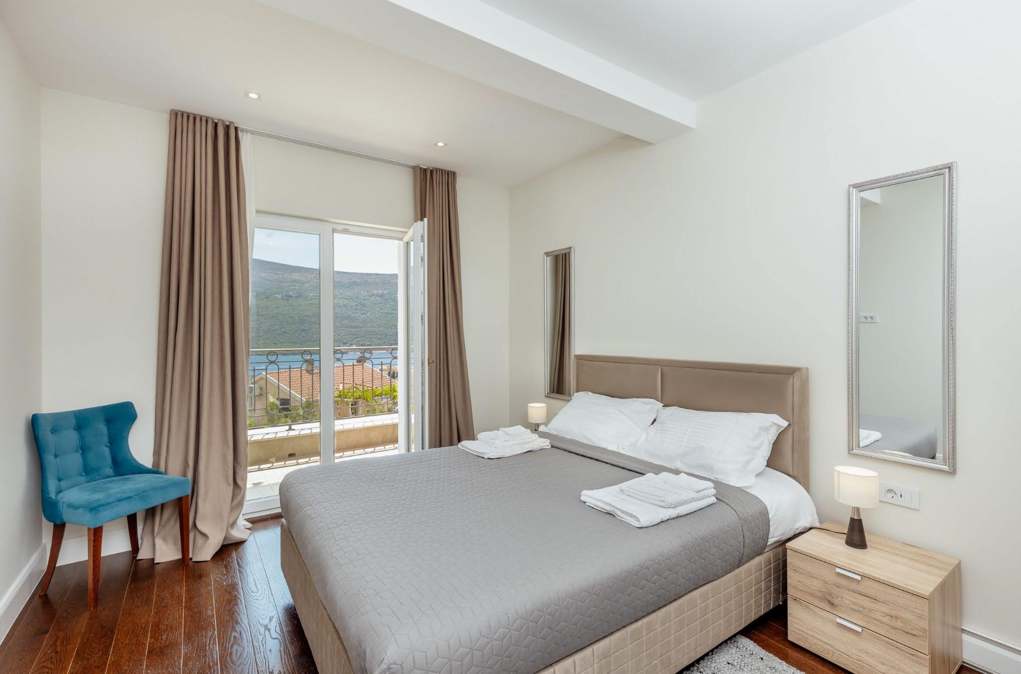 Bedroom in the Kumbor residential complex, Montenegro: a double bed, a blue armchair, and access to a balcony with sea and mountain views.