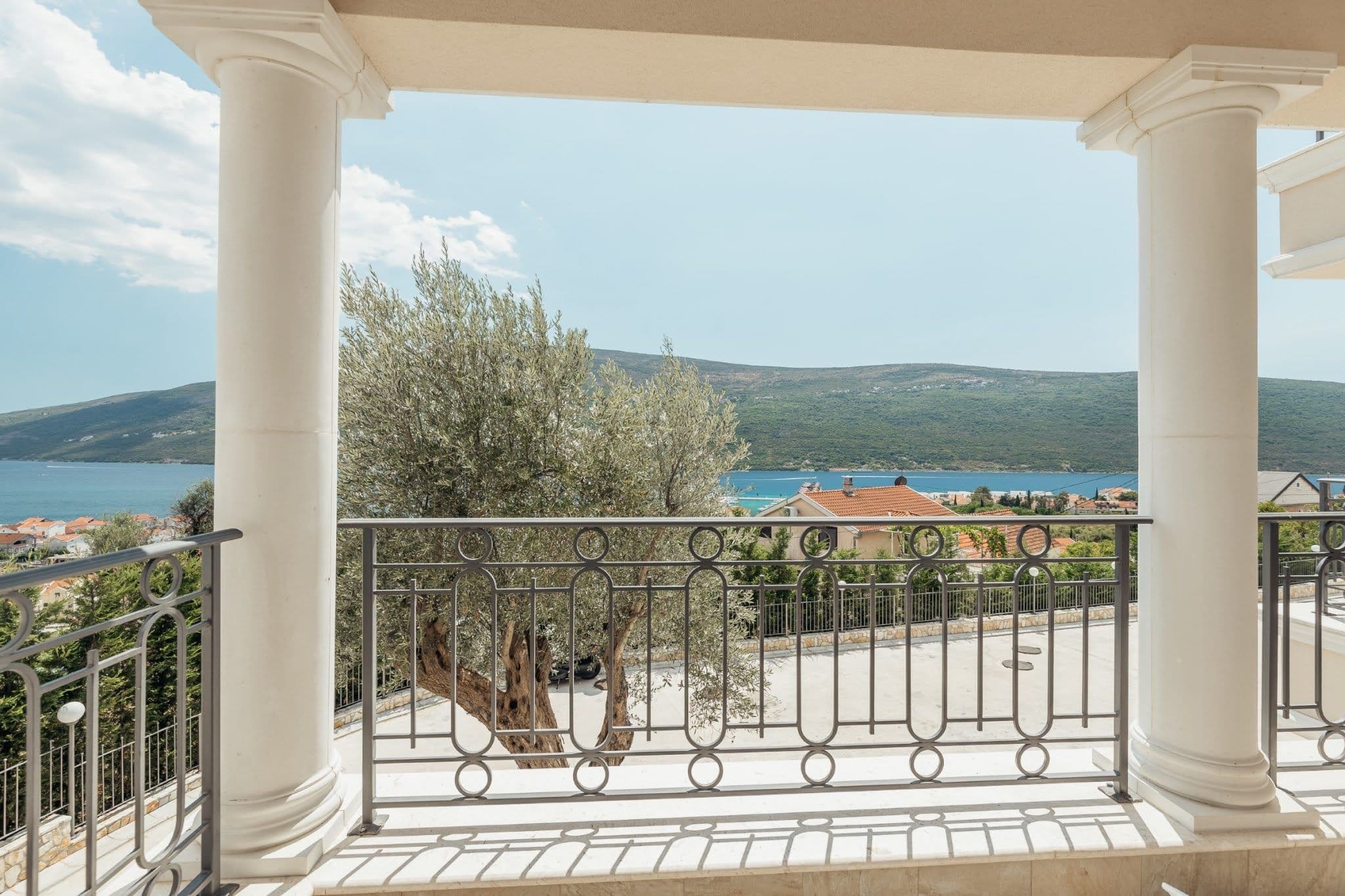 View from a balcony with columns overlooking the sea and mountains in a residential complex, Kumbor, Montenegro. Wrought-iron railings and an olive tree.