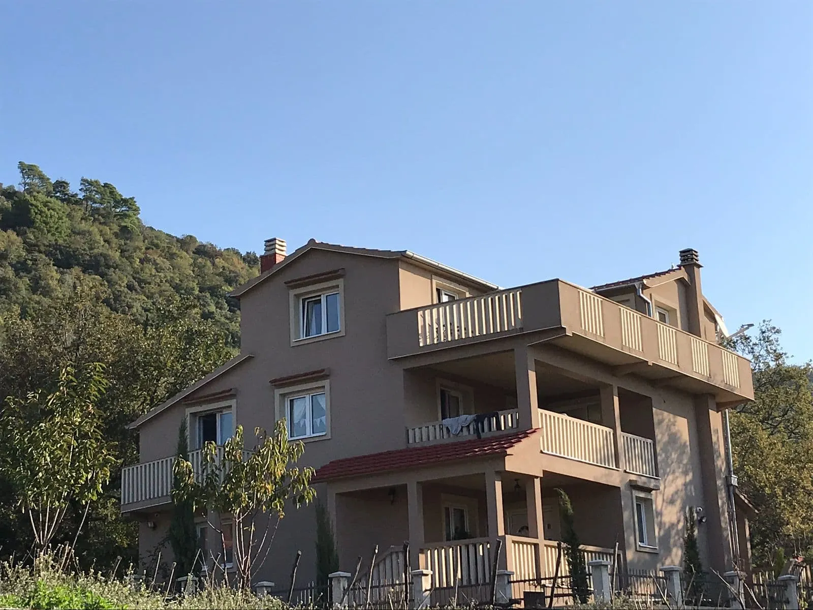 Two-story house with balconies and red tiled roof set against a forested hill under blue sky.