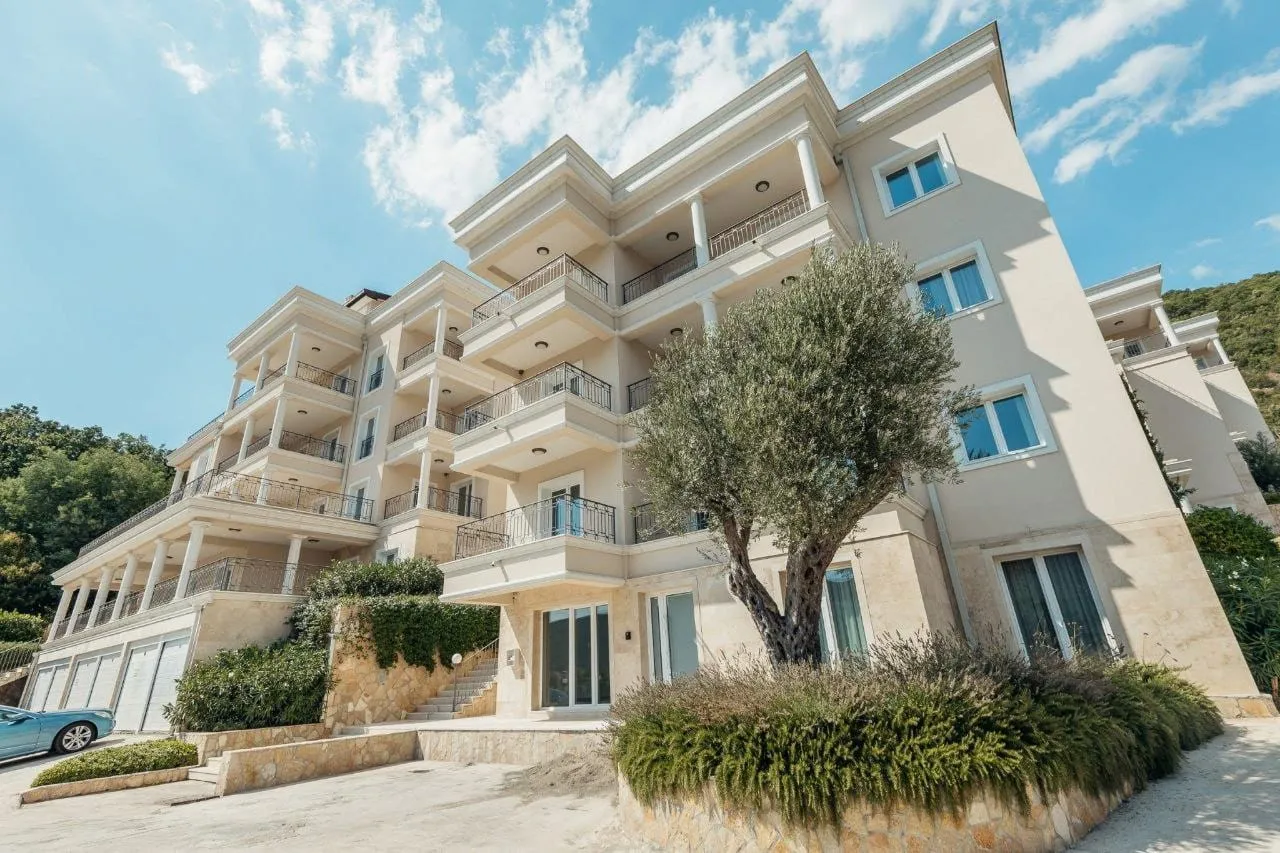 Exterior view of a multi-story residential building featuring balconies, columns, and an olive tree against a blue sky.