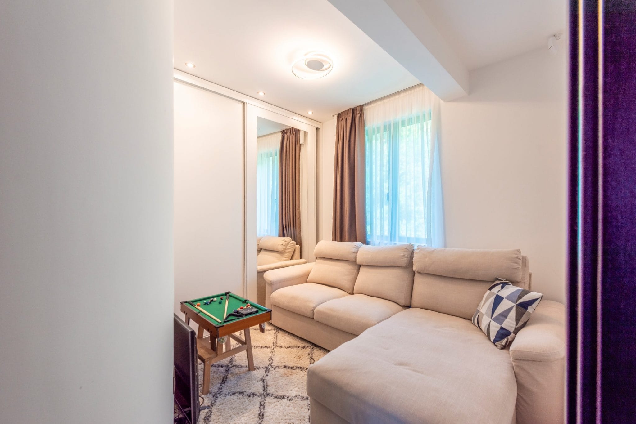 Bright living room with a beige sofa and mirrored wardrobe in a penthouse, Becici, Montenegro. Interior with a rug and mini billiards.