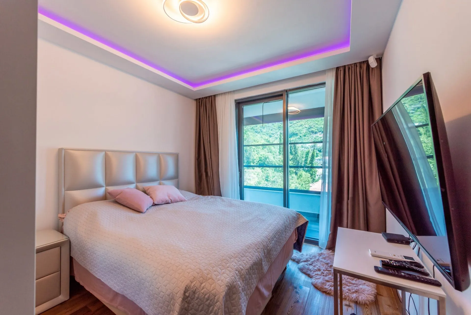 Modern bedroom featuring a double bed with pink pillows, purple ceiling LED strips, and a TV near the balcony door.