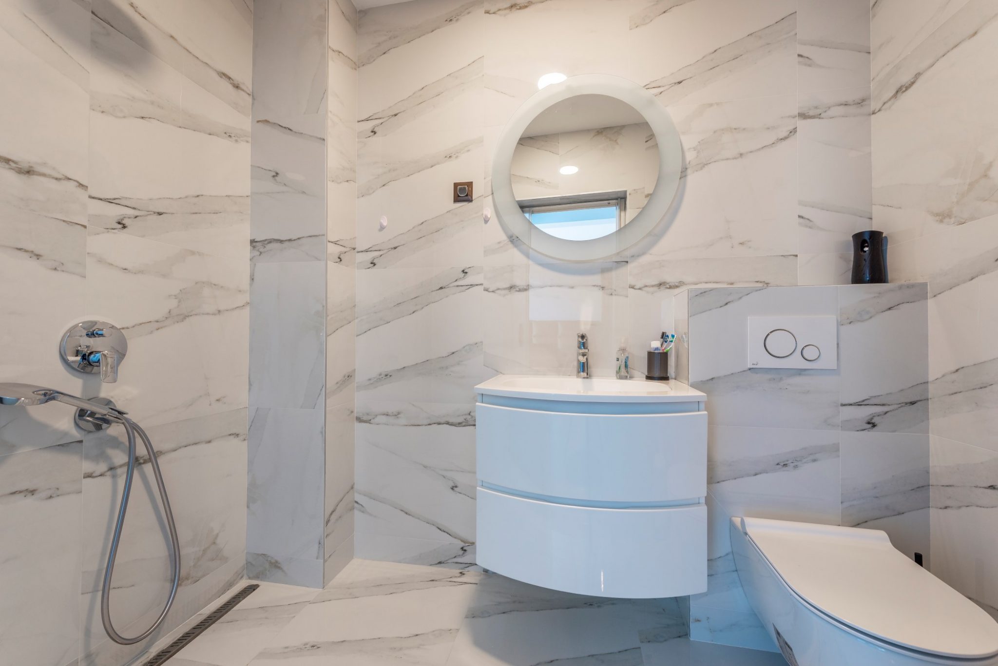 Modern bathroom in a penthouse in Becici, Montenegro: marble-look finish, shower, round mirror, and vanity cabinet.
