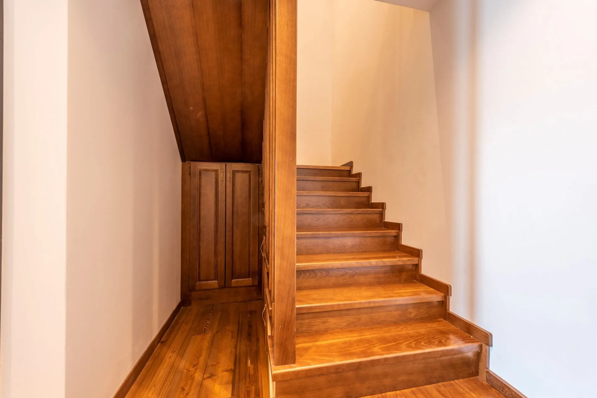 Wooden staircase with built-in cabinets under a sloped ceiling and wooden flooring in a white hallway.
