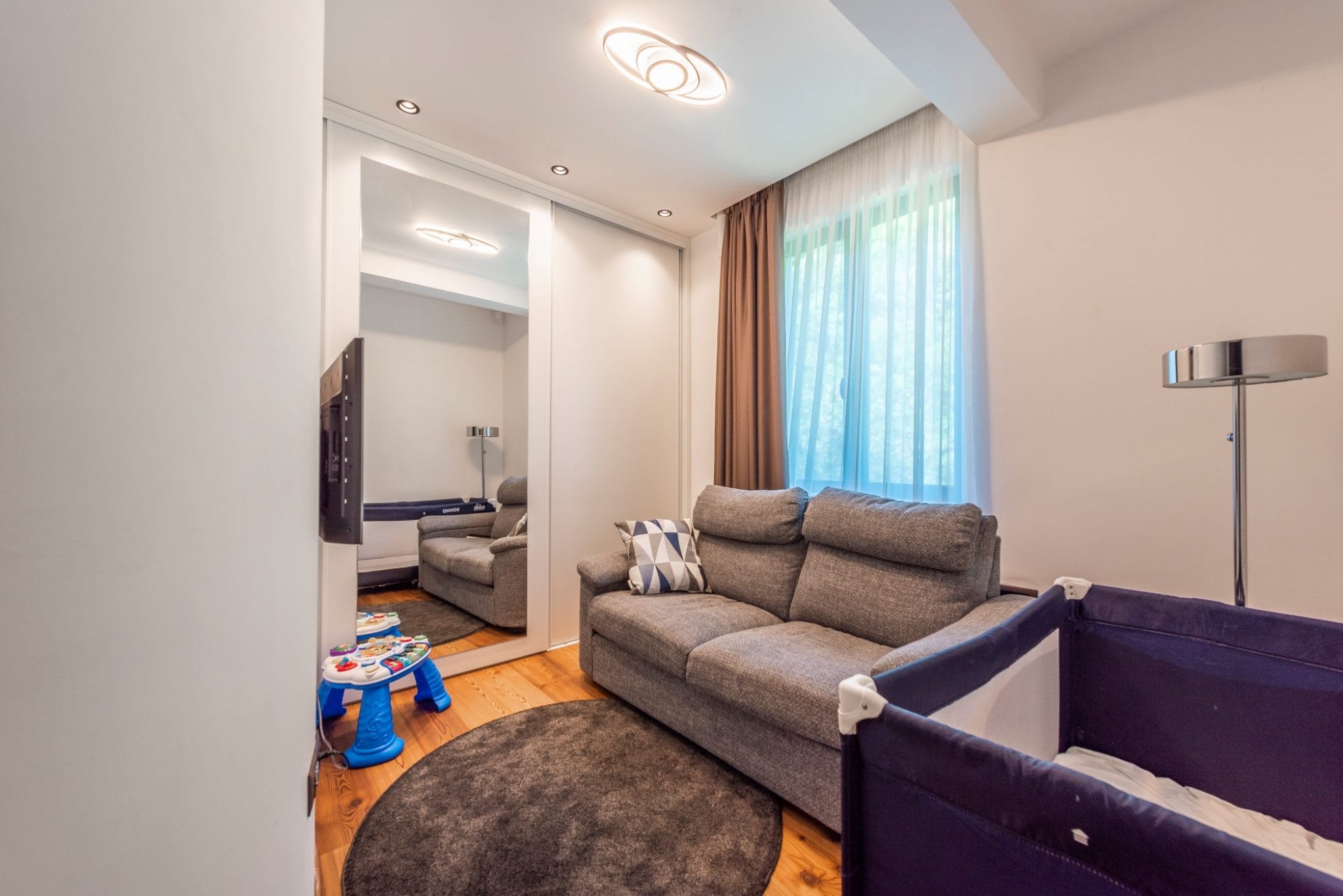Living room in a penthouse in Bečići, Montenegro: gray sofa, mirrored sliding wardrobe, baby crib, and parquet flooring.
