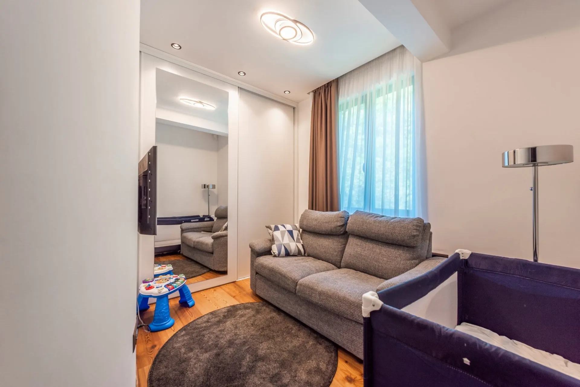Living room featuring a gray sofa, round rug, blue children's activity center, and a purple playpen.