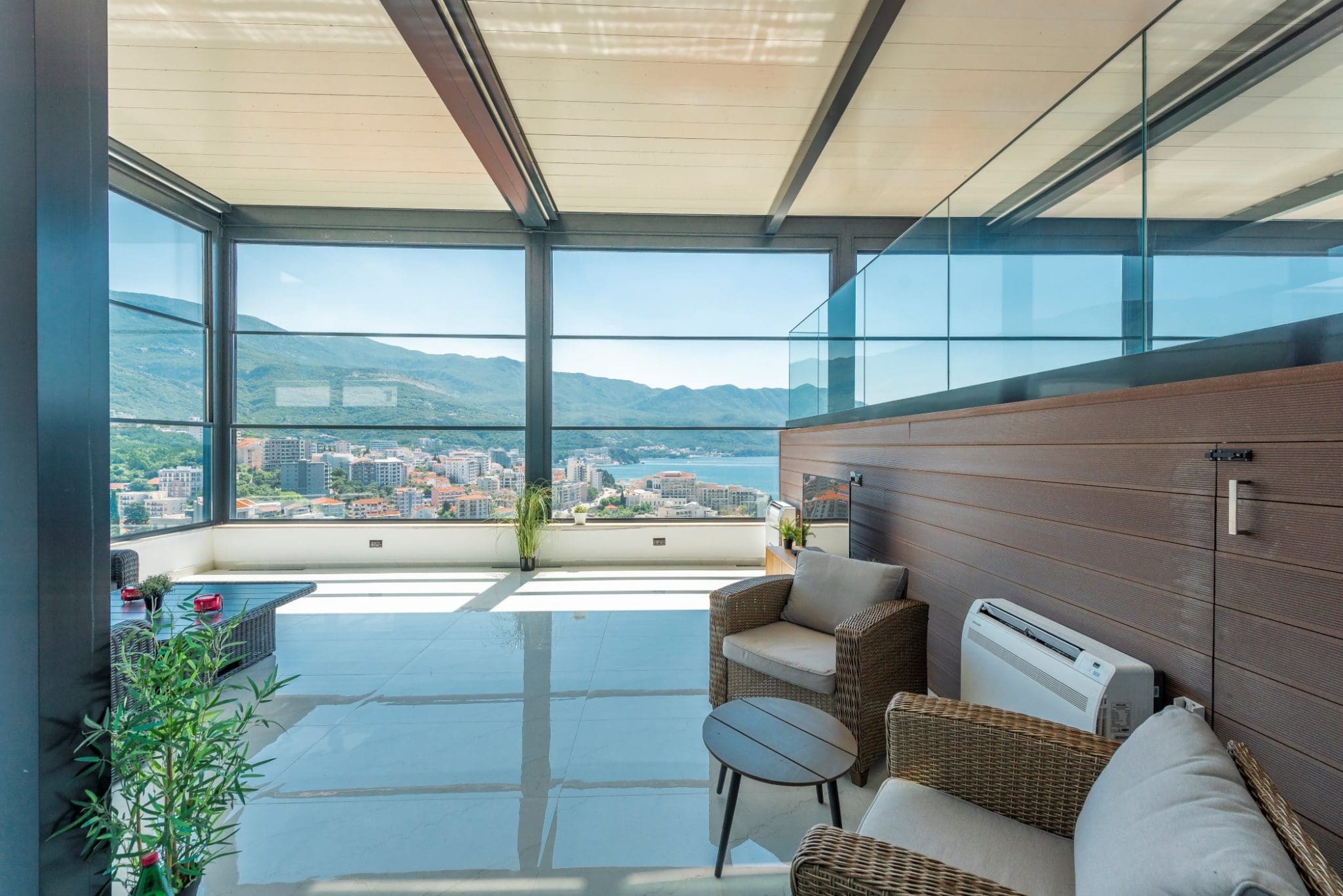 Penthouse terrace in Bečići, Montenegro: panoramic glazing, wicker furniture, and views of the sea and mountains.