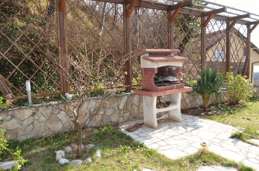 Barbecue area with a stone grill on the apartment terrace in Bar, Montenegro, with a wooden pergola and garden.